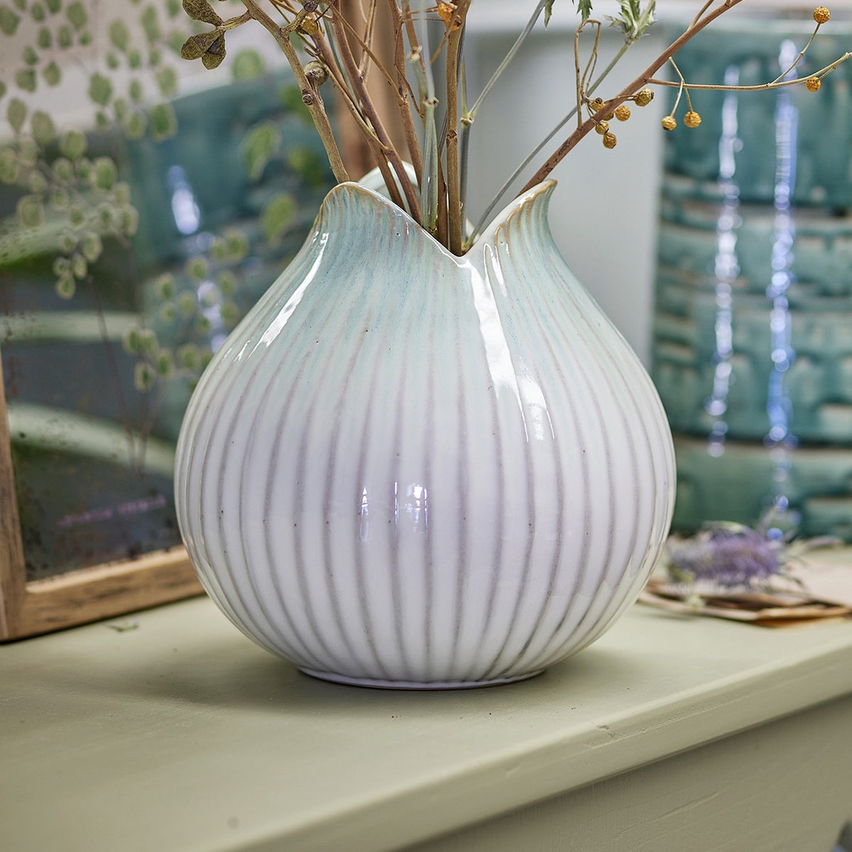 Skylar Fluted Stoneware Vase 19.5cm in white, round ribbed body with a folded petal-like rim, shown holding dried branches on a shelf setting.