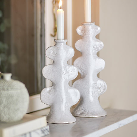 Cason stoneware candle holders in white, a tall and short pair with sculptural rounded lobes, sunburst detailing, and a textured glaze, shown with lit candles on a mantelpiece.
