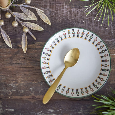 Cream stoneware side plate from the Jolly Christmas set, bordered with a ring of nutcracker figures, holding a gold teaspoon on a dark wood table with festive greenery.