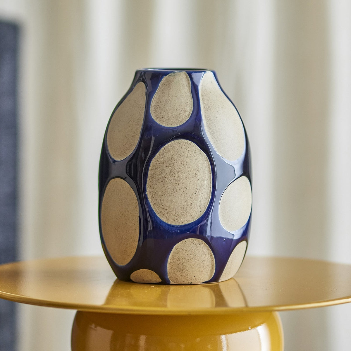Bashir Oval Stoneware Vase 20cm in glossy blue with matt speckled oval pattern, narrow mouth and pebble-like body, displayed on a mustard side table.
