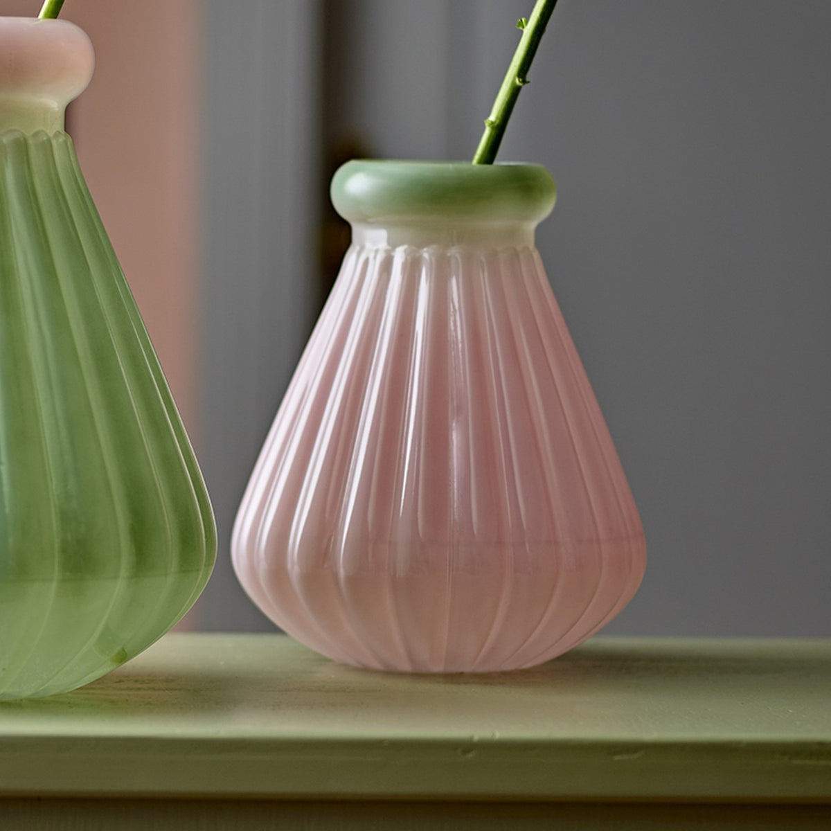 Judith Fluted Glass Vase 20cm in pink, with a narrow green rolled rim and ridged glass body, shown beside a green vase on a light surface.