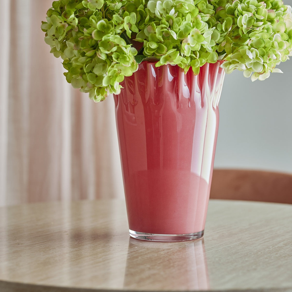Janine Fluted Glass Vase 30cm in pink with wavy, frilled rim and tapered glass body, shown with green hydrangeas on a round table against soft pink curtains.