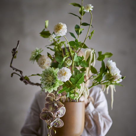 White artificial bouquet in a tan vase with open white blooms, green pompom and dahlia-style heads, orchid sprays, trailing tassels, and a bare branch.