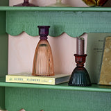 Shilo ribbed glass candle holders in amber and deep purple on a green scalloped shelf, with a matching burgundy holder in dark tones beside books.