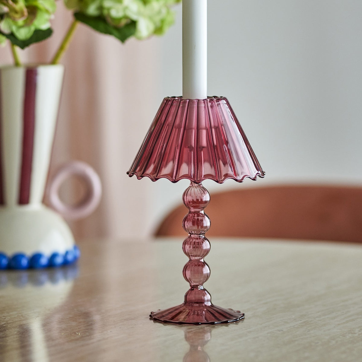 Cisco Glass Tealight Holder in pink, styled as a mini table lamp with a fluted, wavy-rim shade on a stacked bead stem and scalloped foot, shown on a wooden table beside a vase of greenery.