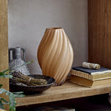 Kanji Glass Vase 30cm in beige with spiral fluted ribs, shown on a wooden shelf beside books, a dark bowl, and a patterned ceramic jug.