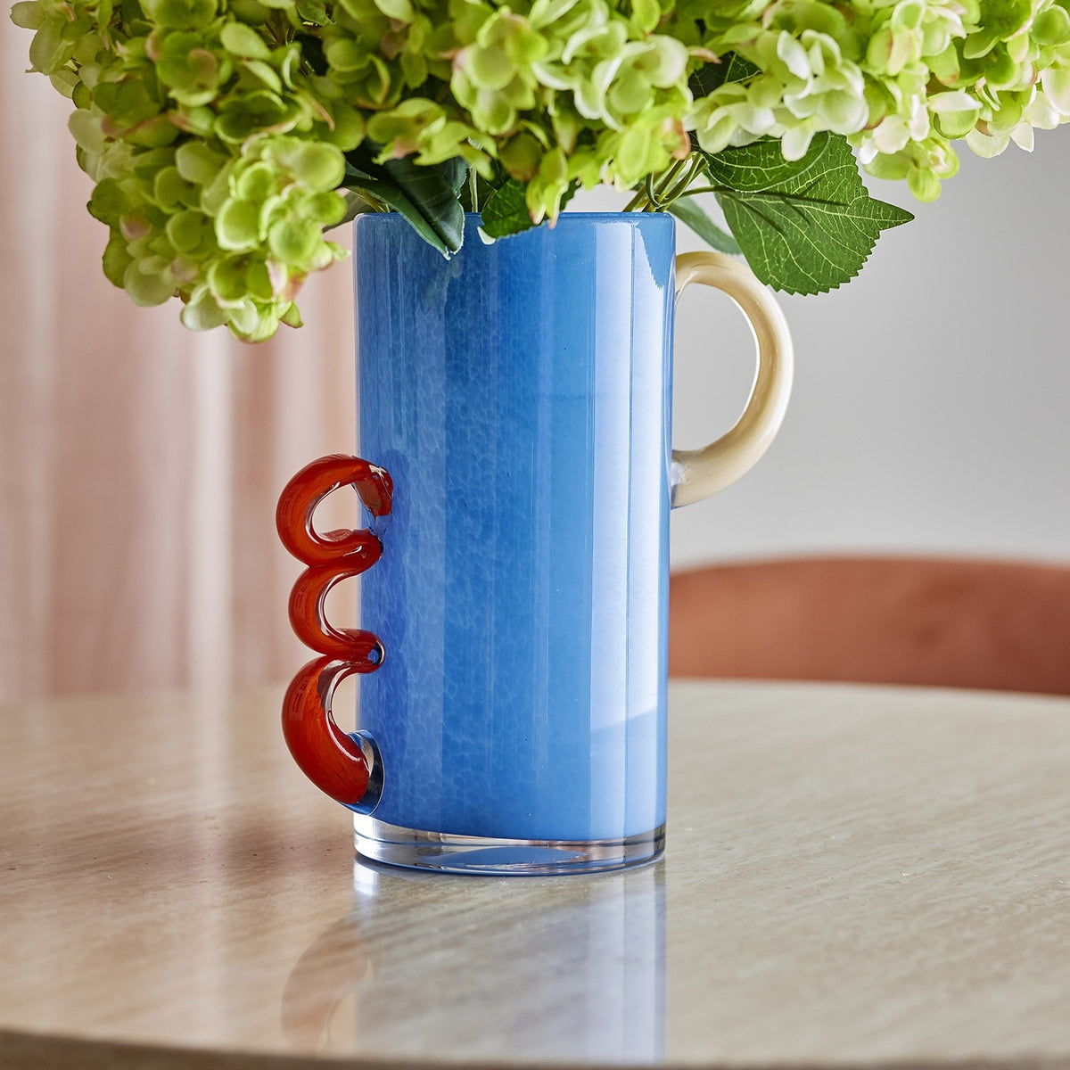 Carlie Glass Vase 25cm in blue with a smooth loop handle and a stacked triple-curl handle in red, holding green hydrangeas on a round table.