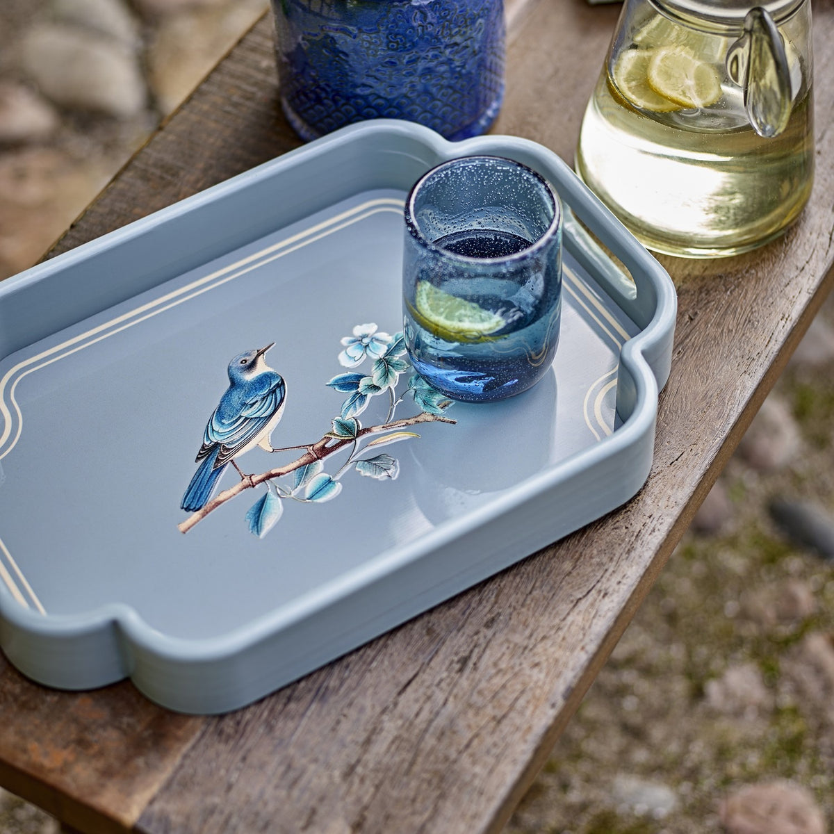Meggie Tray in blue MDF with scalloped rectangular rim and bird-on-branch illustration, shown outdoors on a wooden table beside a glass and jug of lemon water.