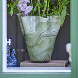 Toryn Marbled Glass Vase 24cm in Green, a wide flared vase with wavy rim, shown holding flowers on a windowsill beside a small blue ornament.