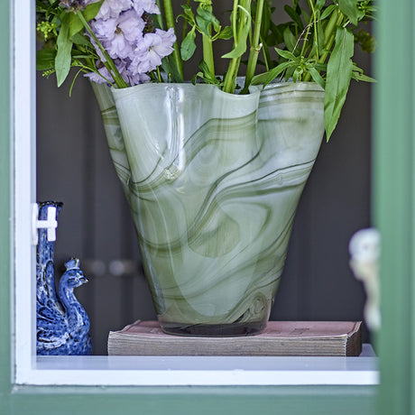 Toryn Marbled Glass Vase 24cm in Green, a wide flared vase with wavy rim, shown holding flowers on a windowsill beside a small blue ornament.