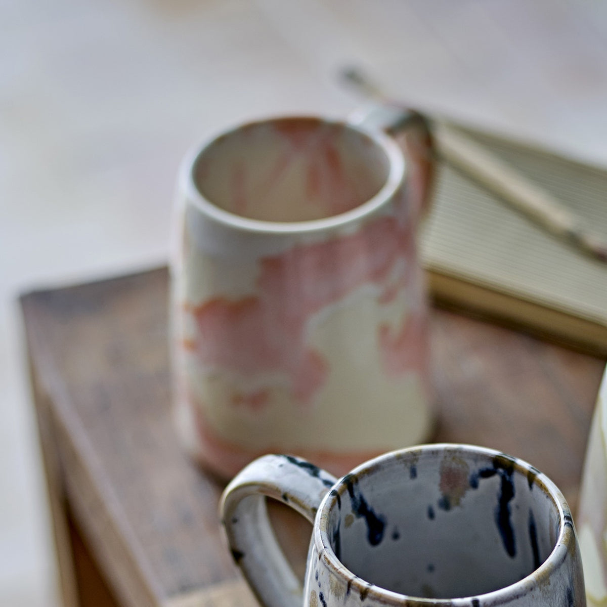 Winnie Marbled Stoneware Mugs in pink-and-cream and grey-and-black reactive glaze finishes, each with a tapered round body and open loop handle, shown on a wooden surface.