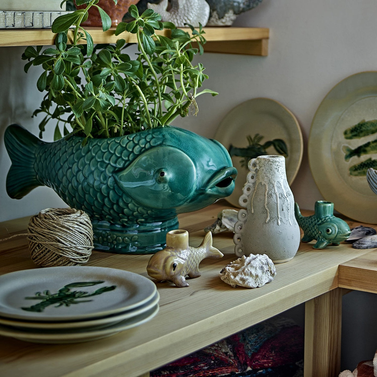 Hiro Fish Stoneware Plant Pot in green reactive glaze with raised scales, shown on a wooden shelf holding a trailing green plant among ceramic decor.