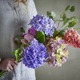 Edge Artificial Bouquet in Purple, held in hand, showing hydrangea-style clusters, pompom heads, and trailing leaves in shades of purple, pink, and green.
