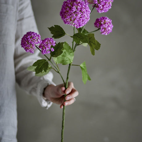 Person in a light linen shirt holding the Snowball Artificial Stem - Purple, showing five pompom-like purple flower heads with serrated green leaves against a plain grey background.