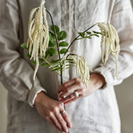 Reed Artificial Stem in cream, held against a linen shirt, showing branching stem with tasselled plumes and rounded green leaves.