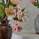 Wild Cherry Artificial Stem in pink, showing ruffled blossoms with stamen centres amid green leaves, arranged in a brown vase beside cream flowers.