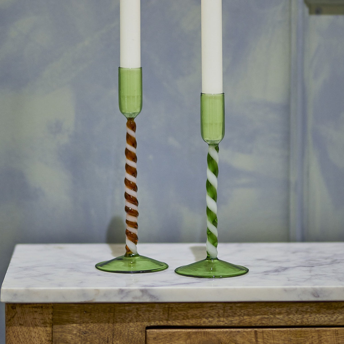 Two mouthblown green glass candle holders with twisted spiral stems, one brown-and-white and one green-and-white, on a marble surface with white taper candles.