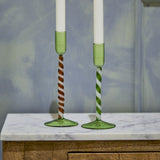 Two mouthblown green glass candle holders with twisted spiral stems, one brown-and-white and one green-and-white, on a marble surface with white taper candles.