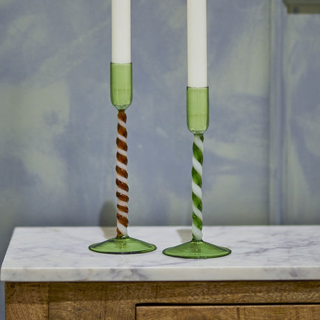 Two mouthblown green glass candle holders with twisted spiral stems, one brown-and-white and one green-and-white, on a marble surface with white taper candles.