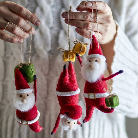 Hands holding the Giles Wool Christmas Tree Decorations Set of 3 in red, felt Santa figures with white beards suspended by twine, shown against a knitted sweater backdrop.