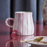 Shirly Striped Stoneware Christmas Mug, white with red vertical stripes and ring handle, holding a teabag, shown on a red surface with a plate and spoon nearby.
