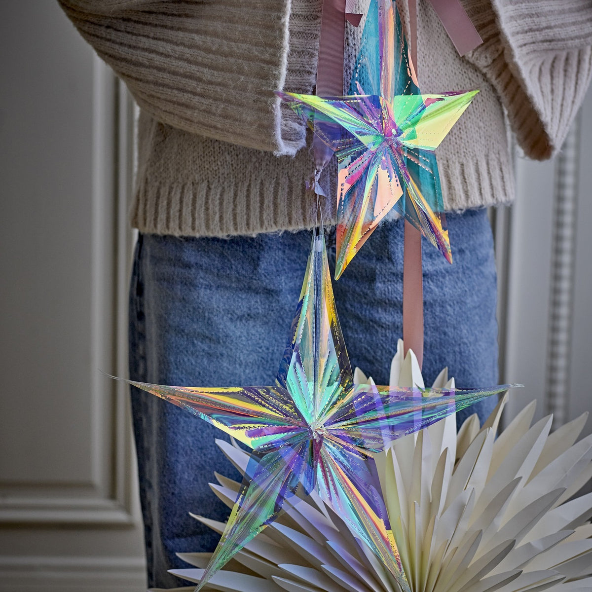 Aikito clear PET star Christmas decorations, set of 2 in different sizes, hung on pink ribbon over a cream jumper and denim skirt.