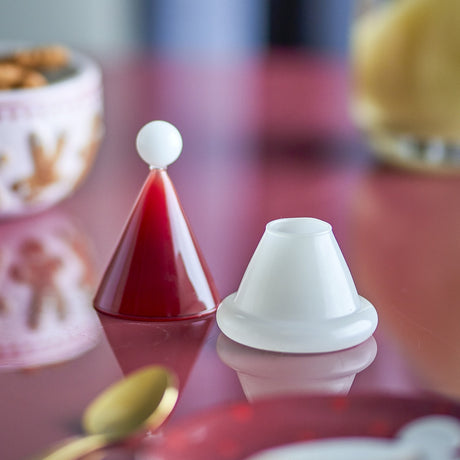 Patrice Glass Christmas Toothpick Holder in red, shaped like a Santa hat with a white ball-tip cone lid and white cup base, shown on a reflective red table.