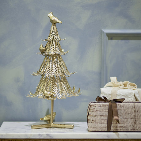 Kiruna Iron Christmas Decoration in gold, a tiered tabletop tree with cut-out leaf patterning and perched birds, displayed on a marble surface beside wrapped gift boxes.