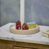 Coze round stoneware candle holder in multicoloured glaze, with three miniature houses in red, green and yellow on a shallow rimmed base around a central taper candle cup, shown on a marble surface.