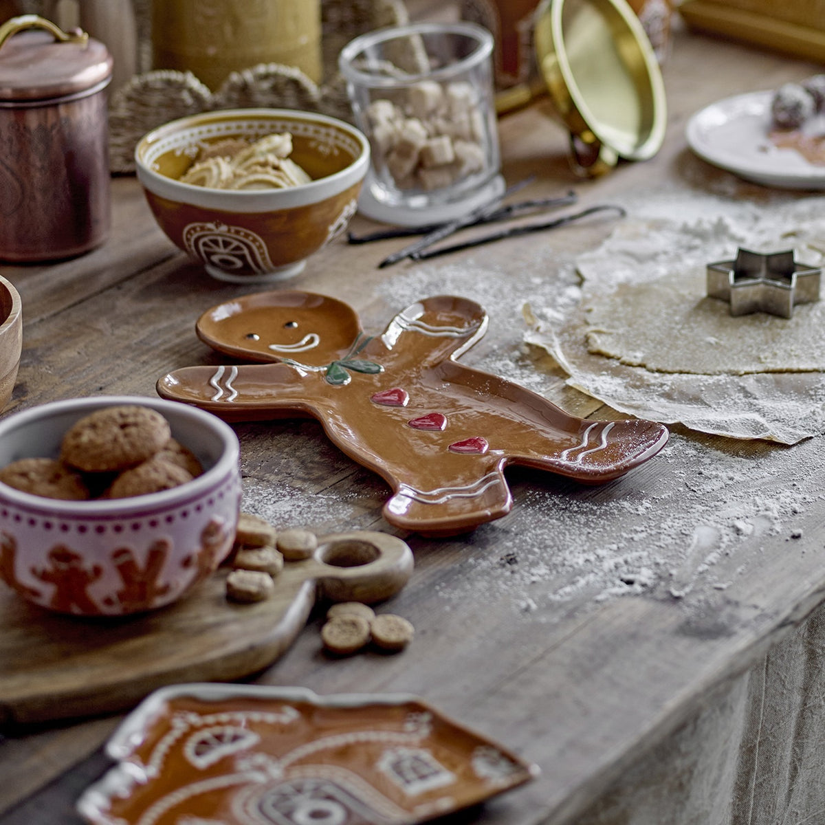 Ginnie Gingerbread Man Stoneware Christmas Platter in glossy brown, shaped like a smiling gingerbread man with a bow, hearts and icing details, shown on a kitchen table amid baking props.
