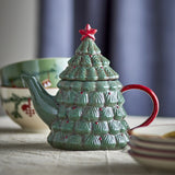 Green ceramic Christmas tree-shaped teapot with layered branch texture, red star knob and red handle, displayed near patterned bowls and plates.