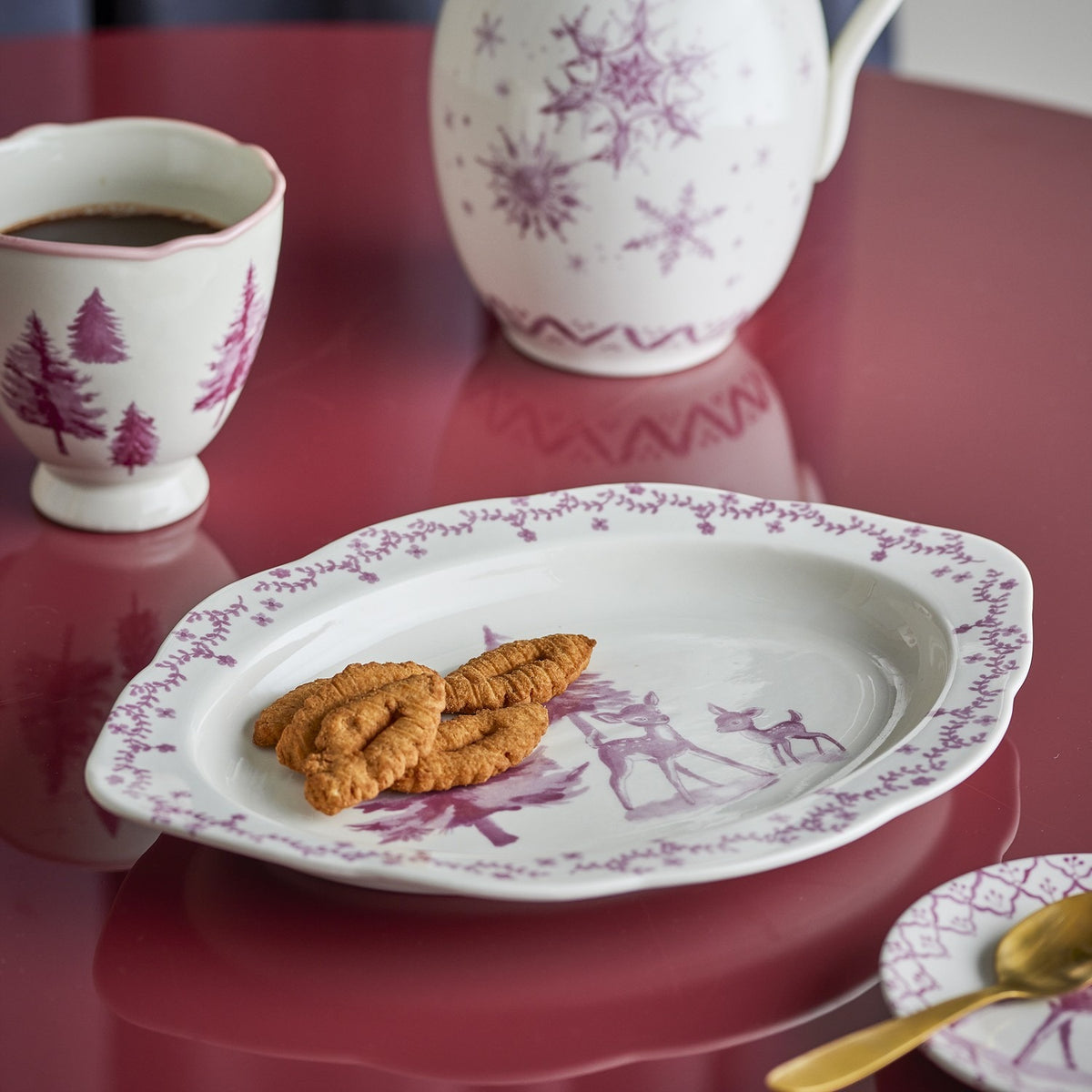 Wonderland Wavy Stoneware Christmas Platter in Pink, oval with scalloped edge, featuring a pink deer and fir tree scene and floral rim, shown with pretzels, a cup, and jug on a red table.