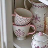 Stoneware Christmas mugs with pink scalloped rims and handles, featuring snowflake and lattice patterns, stacked on a shelf beside matching pink-trimmed jugs.