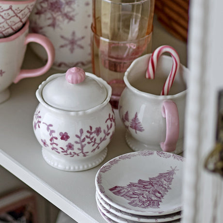Cream ceramic Wonderland milk jug and lidded sugar bowl with pink floral and tree motifs, pink handle and knob, on scalloped beaded feet, displayed on a shelf with pink glassware and patterned plates.