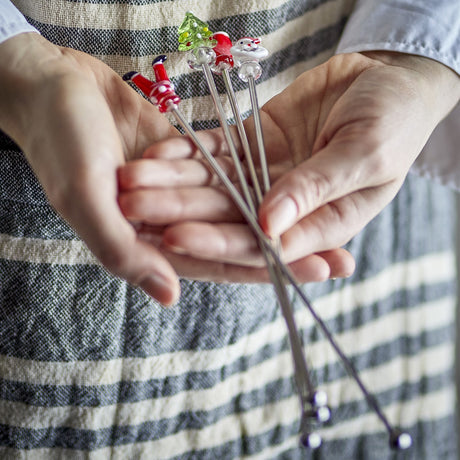 Misa Stainless Steel Christmas Cocktail Stirrers Set of 4 held in hand, showing metal stems topped with glass figures including Santa, a tree, and Santa's legs.