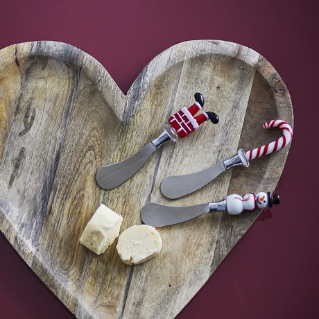 Misa Glass-Handled Christmas Butter Knives, set of 3 with stainless steel blades and festive handles shaped as a candy cane, chimney with legs, and snowman, shown with cheese on a wooden board.