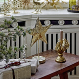 Nilos Star Iron Christmas Decoration in brass, a hammered-texture standing star on a slim stem with round base, displayed on a wooden table beside a gold candlestick and festive décor.