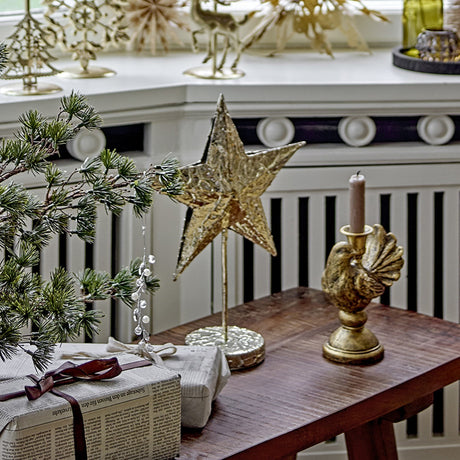 Nilos Star Iron Christmas Decoration in brass, a hammered-texture standing star on a slim stem with round base, displayed on a wooden table beside a gold candlestick and festive décor.