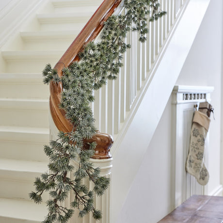 Cedar Artificial Christmas Garland in frosted green trailing over a white staircase banister, with cream stockings visible near a radiator cover below.