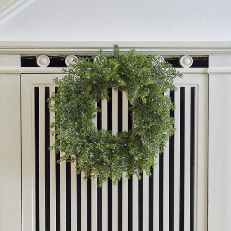 Juniper Artificial Christmas Wreath in green, with full sprigs and small round berries, hung on a cream panelled cabinet with black stripe detailing.