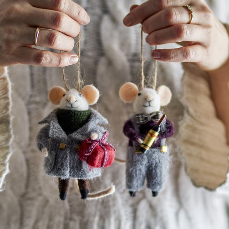 Two hands hold Peo grey wool mouse Christmas decorations by their string loops, one holding a red parcel, the other holding a wine bottle, against a cream knit jumper.
