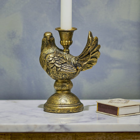Fosna Bird Resin Candle Holder in brass, shaped as a perched bird with carved feathers and fanned tail on a pedestal base, holding a white taper candle on a marble surface.