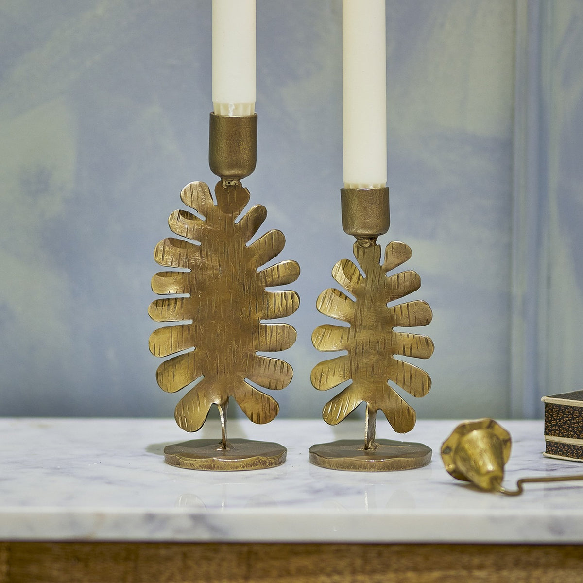 Oaken Iron Candle Holders Set of 2 in gold, each a textured leaf-shaped stand on a slim stem and round base, holding white taper candles on a marble surface.