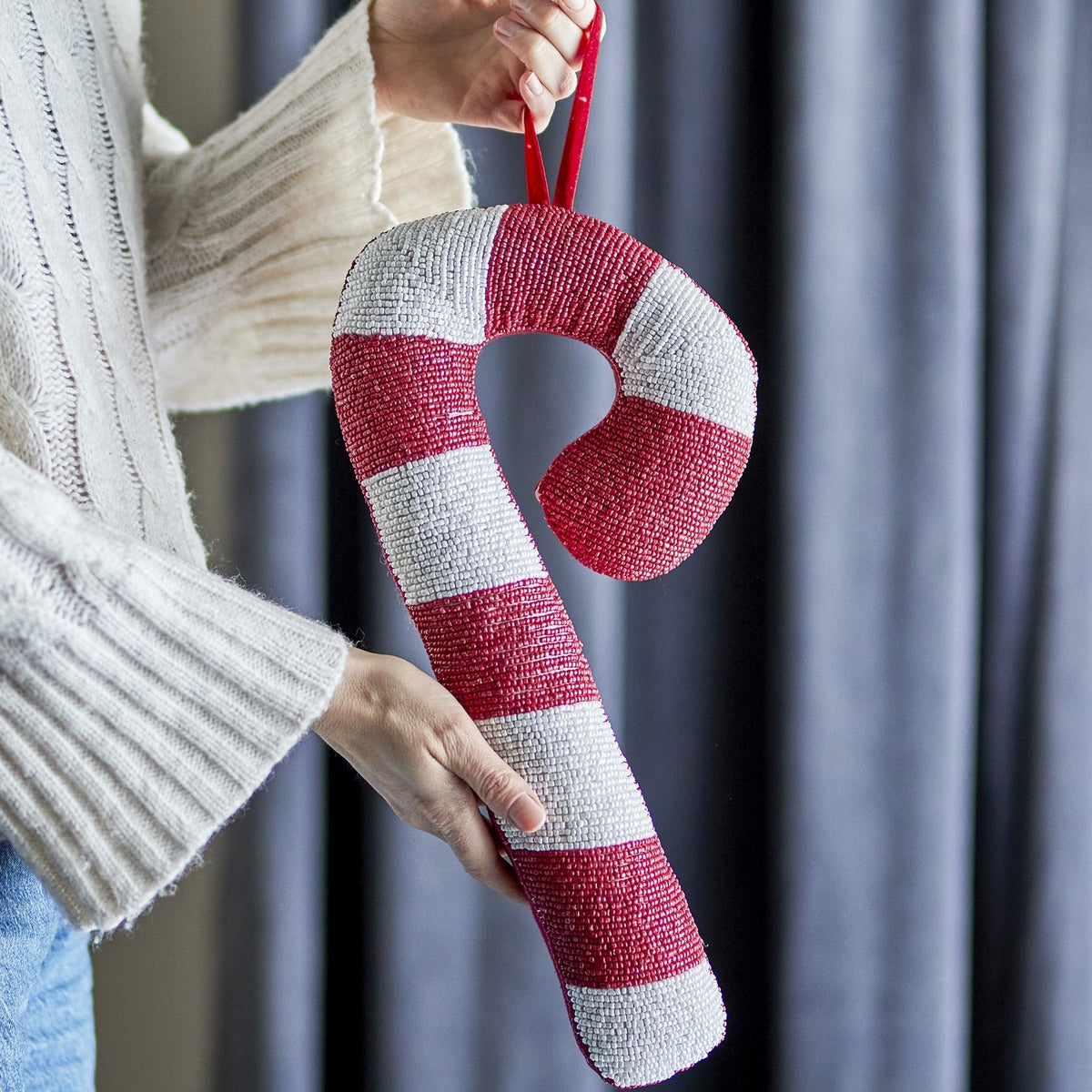 Kunai Striped Christmas Tree Decoration in Red, a padded candy cane-shaped ornament with red and white beaded stripes, held by a person in a knitted jumper against grey curtains.