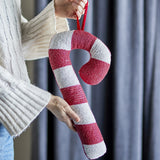 Kunai Striped Christmas Tree Decoration in Red, a padded candy cane-shaped ornament with red and white beaded stripes, held by a person in a knitted jumper against grey curtains.