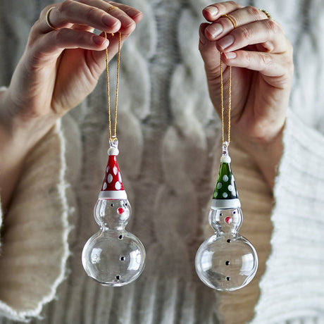 Two hands hold clear glass snowman Christmas decorations by gold cord, each with a painted dotted hat in red or green, against a knitted sweater background.
