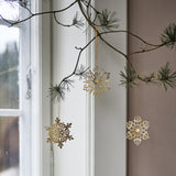 Mani Iron Christmas Tree Decorations Set of 3 in Gold, three pierced metal snowflake ornaments hanging from a pine branch by a window.