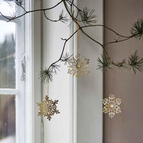 Mani Iron Christmas Tree Decorations Set of 3 in Gold, three pierced metal snowflake ornaments hanging from a pine branch by a window.