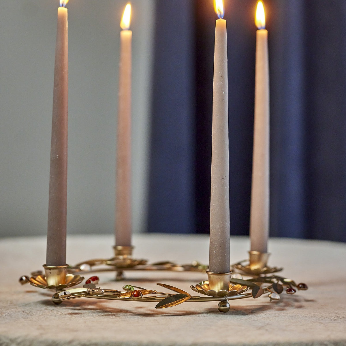 Gold floral iron Advent candle holder with four flower-shaped cups holding lit taper candles, leaf sprigs and coloured bead accents, on a light surface against a dark backdrop.