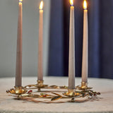 Gold floral iron Advent candle holder with four flower-shaped cups holding lit taper candles, leaf sprigs and coloured bead accents, on a light surface against a dark backdrop.